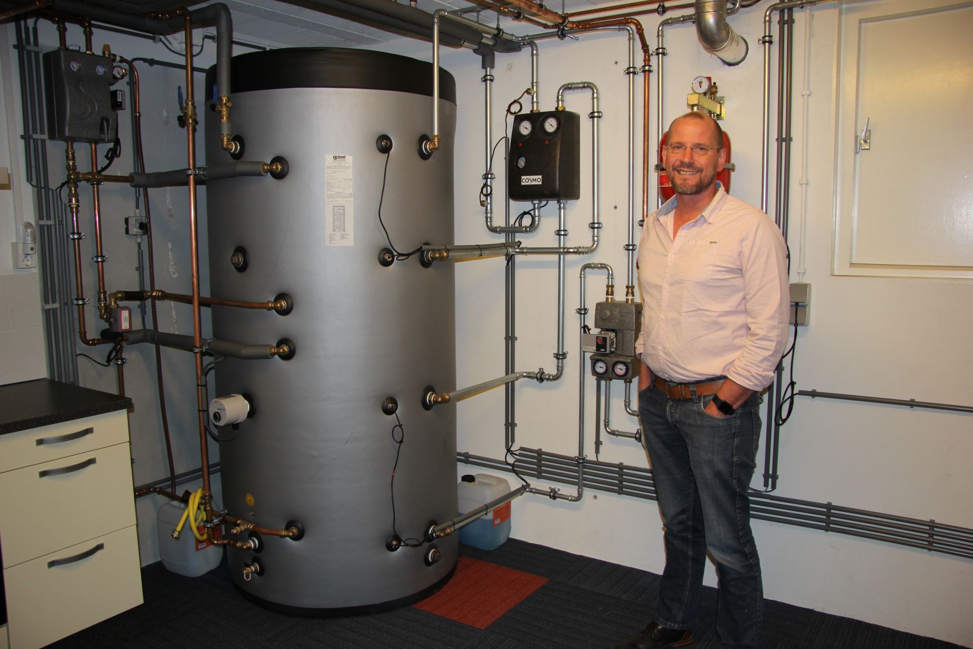 Een man staat trots in de installatieruimte van zijn huis. Er lopen veel leidingen langs de muur en er staat een warmwaterbuffervat.