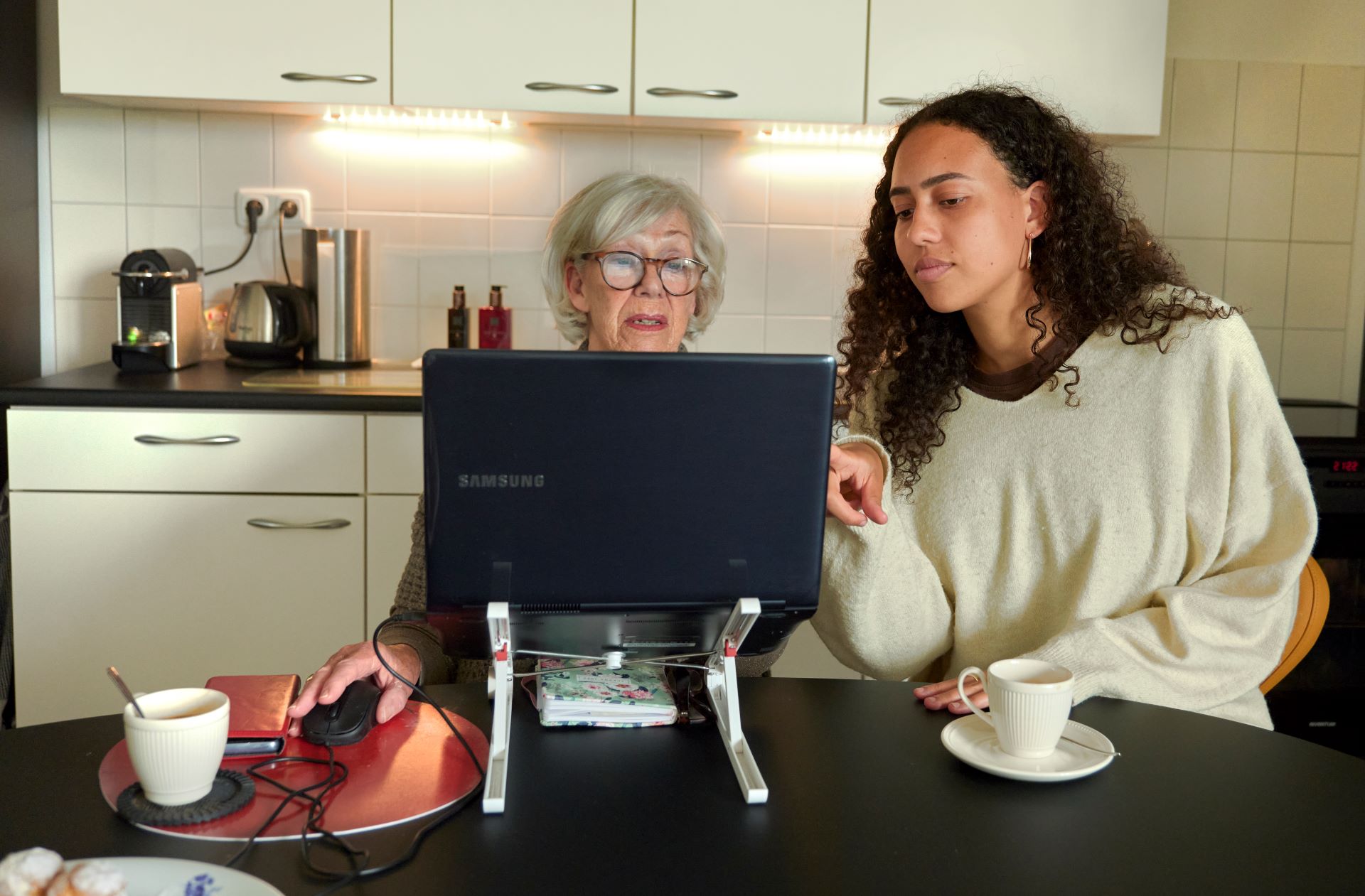 Een jonge en oudere vrouw zitten achter de laptop. De jonge vrouw legt de oudere vrouw wat uit.