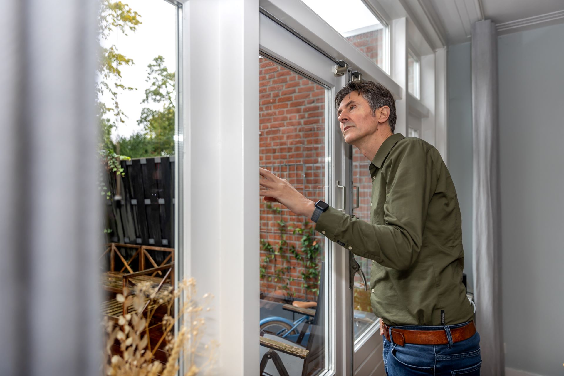 Er staat een man binnen in huis voor het raam. Hij bekijkt de sponning van het raam om te zien wat voor soort glas het is.